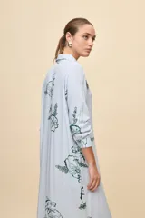 Vestido camisero largo de calce relajado, con patrón de rayas verticales celestes y blancas, y estampa floral en color verde. Posee cuello camisero, mangas largas y cierre frontal con botones.