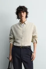 Camisa de algodón de corte relajado, con cuello solapa abotonado (button-down) y manga larga terminada en puño con botón. Presenta un patrón de rayas verticales finas en tonos beige claro y marrón/verde oliva.