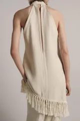 Top halter color crudo, de escote en V y diseño confeccionado en hilatura con mezcla de viscosa. Presenta bajo asimétrico con flecos al tono y cierre en la espalda.
