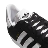 Championes adidas modelo Gazelle, de diseño clásico con exterior de gamuza negra, las icónicas tres tiras laterales en blanco y suela de goma blanca. Presenta el nombre del modelo en letras doradas en el lateral y el logo del trébol en la lengüeta y el talón.