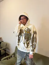 Hoodie oversize de color blanco con estampado artístico en tonos marrones y dorados en el pecho y las mangas. Presenta un forro interior de capucha en tejido marrón texturizado y costuras reforzadas.