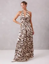 Vestido largo de tejido satinado plisado con estampado animal print tipo cebra en tonos tierra y ocre. Presenta escote halter profundo en V con drapeado central y broche de pedrería, espalda descubierta y falda fluida con apertura frontal.