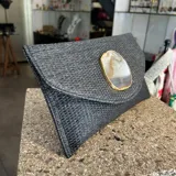 Sobre de fiesta tipo clutch, tejido artesanalmente en la India con rafia color negro grisáceo y aplique de piedra ágata natural con borde dorado. Incluye asa de cadena dorada para colgar al hombro.