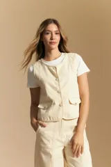 Chaleco de tela color crema, con cuello redondo, cierre frontal de botones y dos bolsillos con tapa en la parte inferior. Presenta costuras contrastantes en color marrón claro.