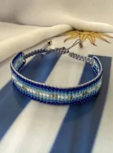 Pulsera tejida con cuentas Miyuki en tonos degradé de azul y celeste, con detalles en blanco y dorado. Cuenta con un sistema de cierre ajustable mediante cordón.