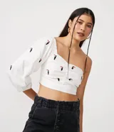 Blusa cropped de lino blanca con manga tres cuartos, hombro asimétrico, escote corazón, lastex en la espalda y bordados de tucanes.