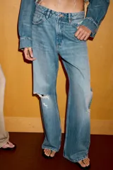 Jeans de corte boyfriend y tiro medio, con diseño de cinco bolsillos, cierre frontal con cremallera y botón metálico. Presenta un acabado lavado con detalles de roturas y desgastes.