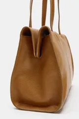 Bolso de hombro de piel color tostado, con detalle de costuras verticales pespunteadas en el cuerpo, doble asa de hombro y cierre con botones imantados.
