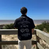Camisa de lino negra con corte boxy, manga corta y cuello clásico. Presenta un estampado gráfico en la espalda con motivos tribales en tonos grisáceos.