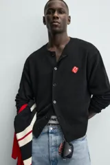 Cárdigan de punto negro, corte relajado, con cuello redondo, manga larga y cierre frontal de botones oscuros. Presenta un parche bordado en forma de dado rojo con corazones blancos en el lado izquierdo del pecho.