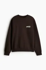Buzo de cuello redondo en color marrón oscuro, con diseño de hombros caídos y estampado pequeño en el pecho con la frase 'Leave Only Footprints'. Presenta puños y bajo acanalados.