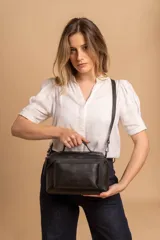 Bandolera de diseño rectangular en color suela, confeccionada en material sintético. Cuenta con un asa superior fija, correa larga ajustable y extraíble, y dos bolsillos frontales con cierre metálico, uno de ellos con diseño superpuesto. Presenta el logo de la marca grabado en el frente.