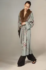 Kimono de largo midi en color verde grisáceo, con cuello de solapa en pelo sintético marrón. Presenta mangas largas acampanadas, cierre frontal con cinturón ajustable del mismo tejido y un delicado bordado floral en toda la prenda.