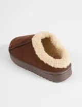 Pantufla tipo mocasín color marrón, con exterior de nylon y forro interior de corderito sintético color crema. Cuenta con una suela de goma dentada de plataforma.