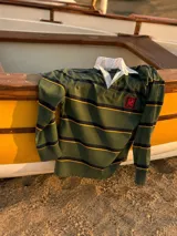 Buzo tipo rugby de manga larga, confeccionado en algodón con diseño de franjas horizontales en verde militar, negro y amarillo. Presenta cuello tipo polo en color blanco y un detalle de bordado rojo en el pecho.
