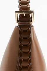 Cartera tipo hobo de diseño estructurado en color marrón, con costuras visibles en contraste y correa de hombro trenzada en tonos marrón y negro con hebilla metálica lateral.