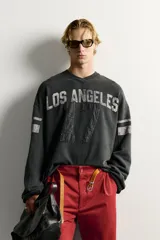 Sudadera cropped fit de color negro con efecto lavado vintage, cuello en V y manga larga. Presenta estampado con texto "LOS ANGELES" y el número "17" en parches acolchados en el frente, además de bandas grises en las mangas.