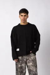 Sweater negro de calce relajado con diseño de textura combinada, presentando un lado liso y otro con tejido de ochos. Cuenta con cuello redondo y una etiqueta decorativa en el frente.