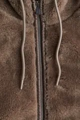 Abrigo de tejido suave efecto peluche en color marrón, con capucha ajustable mediante cordones, cierre frontal de cremallera y bolsillos laterales insertados. Presenta ribetes en los bordes y cuello elevado.