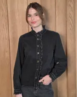Camisa de denim negro con volados en el cuello alto y en la botonera frontal, manga larga con puños fruncidos y cierre de botones metálicos dorados.