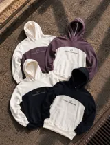 Canguro con capucha y bolsillo tipo canguro, diseño en bloque de color blanco en la parte superior y mangas, y morado oscuro en la parte central e inferior. Presenta el logo de la marca bordado en el pecho.