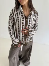 Camisa blanca de manga larga con delicados bordados florales verticales y detalles de encaje. Presenta cuello clásico, cierre frontal con botones y terminaciones festoneadas en los puños y el ruedo.