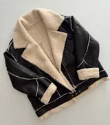 Campera tipo gamulán de cuero sintético negro con interior y detalles de corderito sintético en color crema. Presenta cierre frontal, bolsillos laterales con cierre y cinturón ajustable en el ruedo.