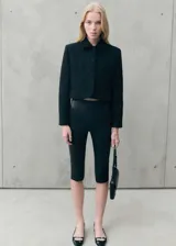 Chaqueta corta de tejido tweed en color negro, con cuello camisero, manga larga y cierre frontal con botones forrados. Presenta un diseño recto con dos bolsillos laterales.