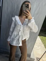 Camisa blanca de tela bordada con diseño floral, cuello en V con volados y detalles de encaje, manga larga con puños elásticos.