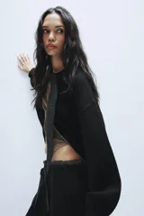 Chaqueta de punto negra con cuello redondo, cierre frontal con cremallera metálica y detalle de lazada en tejido a cuadros en contraste. Presenta mangas largas abullonadas con puños elásticos y bajo elástico.