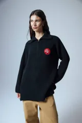 Sweater negro de cuello polo tejido con cierre diente de perro a tono y tajos laterales. Tiene un parche bordado con elementos del drop.