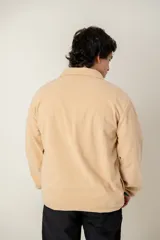 Buzo de pana color beige, de corte oversize, con cuello alto y cierre frontal parcial con botones a presión. Presenta detalles en color verde en la tapeta de los botones y en los puños, además de un parche decorativo en el pecho.