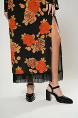 Vestido largo negro con estampado de flores naranjas y amarillas, cuello mao con abertura en forma de gota, mangas largas acampanadas con botones en los puños, tajo lateral y flecos en el ruedo.