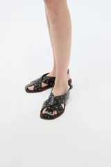 Sandalias de eco cuero negro con tiras cruzadas en el empeine, decoradas con tachas doradas. Cuentan con tira ajustable en el talón con hebilla y suela plana marrón.