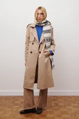 Trench coat color beige con cuello de solapa, cierre frontal abotonado, bolsillos laterales y mangas largas con cintas ajustables. Incluye capucha desmontable y forro interior rayado. La tela exterior cuenta con protección repelente a la lluvia y manchas.