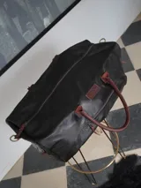 Bolso de viaje tipo weekender confeccionado en cuero negro, con manijas y correa ajustable en cuero color bordó. Presenta cierre metálico frontal con detalle en azul, bolsillo externo con cierre y parche de marca grabado en el frente.