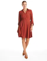 Vestido corto color terracota, confeccionado en crepe, con cuello camisero, escote en V y cierre frontal parcial. Presenta bolsillos decorativos con cierres metálicos en el pecho, mangas largas regulables con presillas y cintura elastizada.