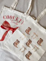 Tote bag de tela tipo lienzo color crudo con estampado frontal. Un modelo presenta un lazo rojo grande con la inscripción 'COQUETTE Girly' y el otro modelo presenta un estampado repetitivo de botas vaqueras y lazos pequeños en tonos beige.