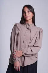Camisa de color azul con cuello alto decorado con volados y lazo de ajuste. Presenta tablas decorativas en los hombros, cierre frontal con botones de nácar y mangas largas con puños elásticos que generan un efecto abullonado.
