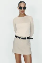 Falda pantalón corta de tiro medio en color beige, con bolsillos delanteros y cierre frontal de cremallera. Incluye un cinturón negro integrado con hebilla metálica redonda.
