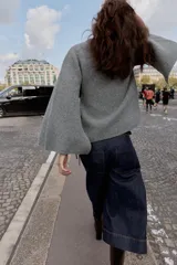 Chaqueta de punto gris con cuello redondo, manga larga acampanada y cierre frontal cruzado con botón y lazada lateral.