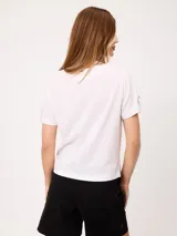 Remera blanca de manga corta con vuelta en los puños y detalle de botón dorado. Presenta cuello redondo y corte recto.