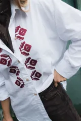 Camisa de algodón blanca con corte clásico y bordado de flores en punto cruz color bordó en la parte frontal. Presenta cuello camisero, cierre frontal con botones y mangas largas con puños.