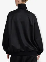 Chaqueta de estilo deportivo y corte oversize, color negro con franjas blancas en las mangas y cuello alto. Cuenta con cierre frontal completo de cremallera y bolsillos laterales.