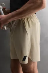 Pantalón corto deportivo color marrón oliva, confeccionado en tejido técnico ligero y elástico, con forro interior. Presenta cintura elástica ajustable con cordón, bolsillos frontales, bolsillo de seguridad trasero con cierre y aberturas laterales en el bajo.