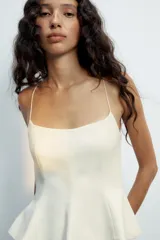 Top blanco de tejido elástico con escote redondo, tirantes finos y bajo con vuelo tipo peplum. Cierre lateral con cremallera oculta.