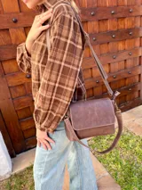 Cartera bandolera de cuero color chocolate con solapa frontal, costuras a la vista y herrajes metálicos. Incluye dos correas desmontables, una corta y una larga, y cuenta con bolsillo interior.