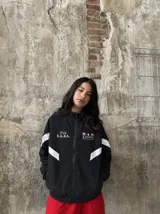 Campera deportiva negra con cuello alto, cierre frontal completo y franjas blancas en las mangas. Presenta estampados en color rosado en el pecho, incluyendo la inscripción "Only F.A.N.S." y gráficos de palmeras con estrellas.