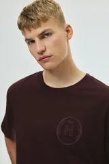 Remera de punto de algodón de peso medio color bordó, con cuello redondo ribeteado, hombros caídos y bajo recto. Presenta un diseño estampado circular en el pecho con texto y un arco. Corte holgado.