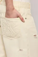 Pantalon de mezclilla color marfil con diseño cruzado asimétrico en la cintura, perneras anchas de corte relajado y detalles de roturas sutiles en la rodilla y el bolsillo.